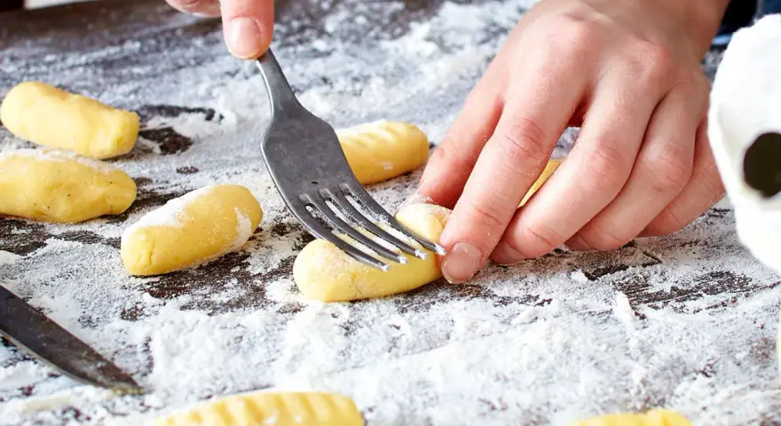 Hände formen mit einer Gabel die Rillen für original Gnocchi auf einer bemehlten Arbeitsfläche.