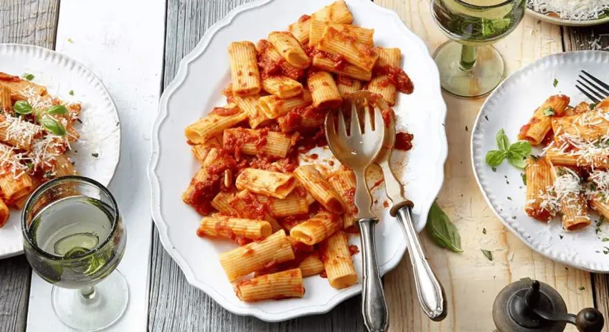 Ein Teller mit Rigatoni mit Tomatensauce, garniert mit Parmesan und Basilikum. Daneben ein Glas Weißwein.