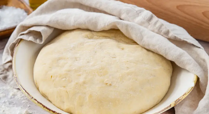 Frisch zubereiteter pizzateig mit trockenhefe ruht unter einem Tuch. Daneben liegen Salz und Olivenöl.