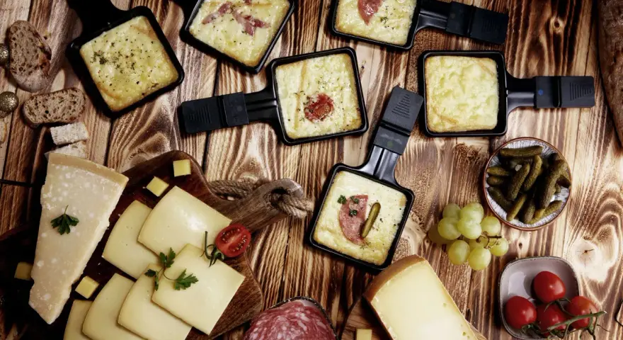 Raclette-Pfännchen mit geschmolzenem Käse, dazu Brot, Käse, Salami, Cornichons und Trauben. Perfekt für einen gemütlichen Abend mit **Wein zum Raclette**.
