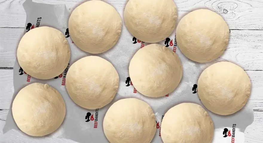 Neun runde, helle pizza teiglinge liegen auf Backpapier mit dem Logo "Beefbandits" auf einer Holzoberfläche.