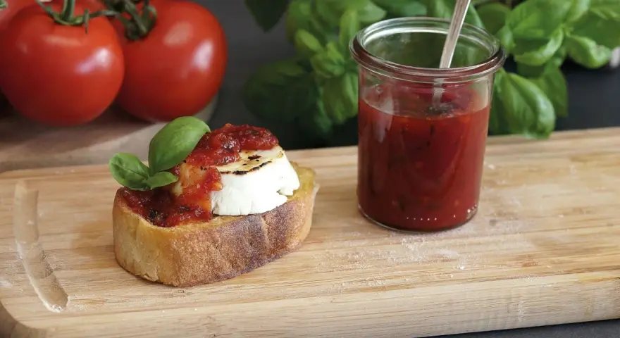 Bruschetta mit Ziegenkäse und Tomatenmarmelade, inspiriert von einem italienischen Tomatenmarmelade Rezept. Frische Tomaten und Basilikum im Hintergrund.