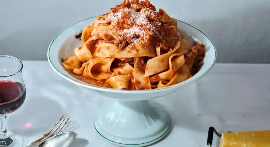 Ein Teller breiter Bandnudeln mit herzhaftem al ragu, bestreut mit Parmesan. Daneben ein Glas Rotwein und Besteck.