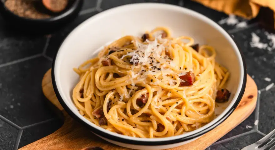 Eine Schüssel mit echter Carbonara, garniert mit geriebenem Parmesan und schwarzem Pfeffer. Daneben ein Holzlöffel mit Pfefferkörnern.