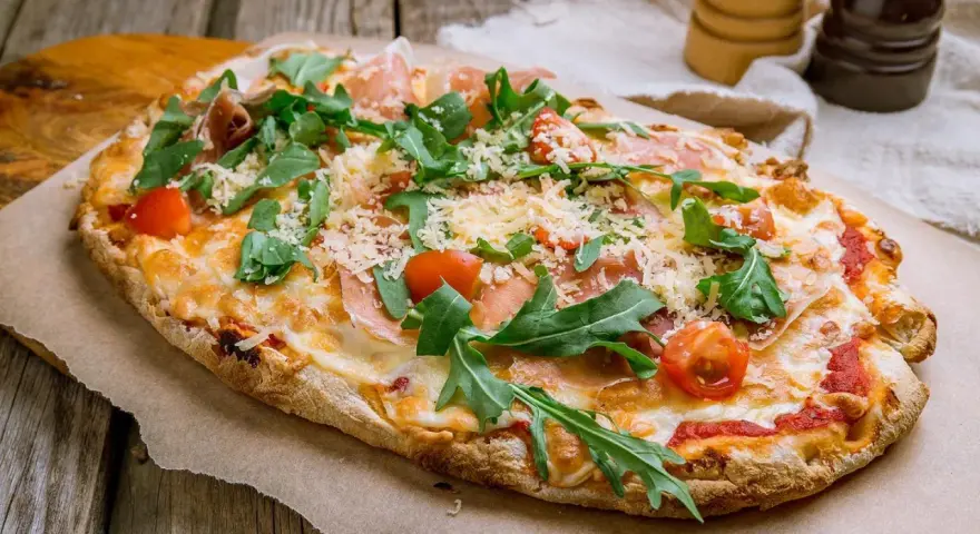 Eine köstliche Pinsa belegt mit Schinken, Rucola, Kirschtomaten und viel Käse auf rustikalem Holzuntergrund.