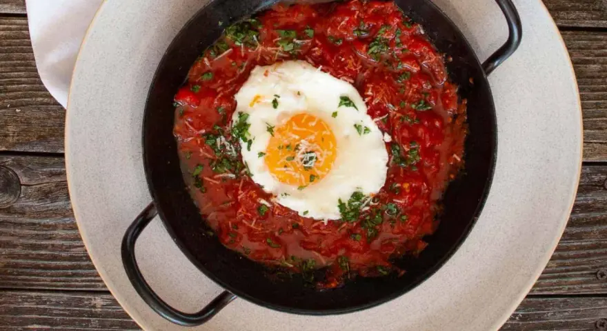 Ein Spiegelei ruht in einer würzigen Tomatensoße, garniert mit Kräutern und Käse. Ein Gericht, das an "uova in purgatorio" erinnert.