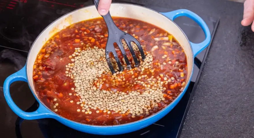 Eine Person rührt Linsen in eine köchelnde Bolognese-Sauce in einem blauen Topf.