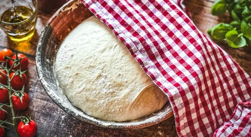 Frischer Pizzateig ruht in einer Schüssel, bedeckt mit einem rot-weiß karierten Tuch. Daneben liegen Kirschtomaten und ein Glas Olivenöl – die perfekten pizzateig zutaten.