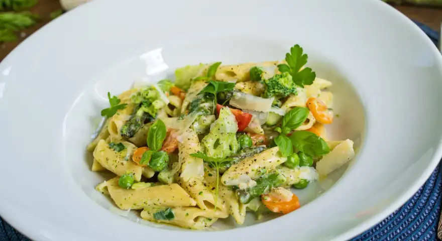 Penne mit cremigem Pesto, Spargel, Erbsen, Brokkoli und Karotten, garniert mit Parmesan und frischen Kräutern.