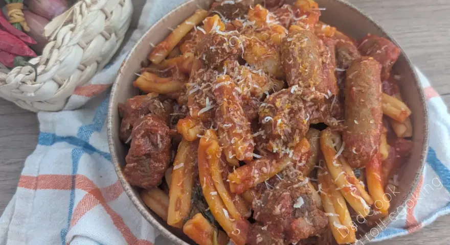 Herzhaftes Pasta Ragu mit Fleischstücken und geriebenem Käse, serviert in einer Schüssel auf einem karierten Tuch. Daneben ein Korb mit Knoblauch, Schalotten und Chilis.