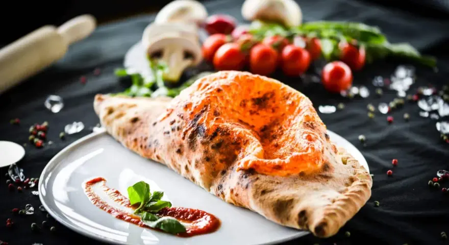 Ein goldbrauner, knuspriger Pizza Calzone mit geschmolzenem Käse und frischem Basilikum auf einem Teller.