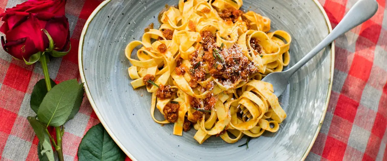 Ein Teller Tagliatelle mit herzhaftem rag&ugrave; alla bolognese, garniert mit Parmesan und Rosmarin. Eine rote Rose liegt daneben.
