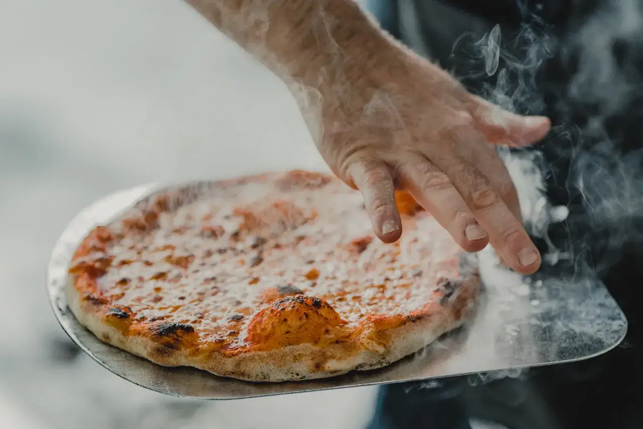 Eine Hand schiebt eine frisch gebackene Pizza mit dampfendem pizzateig auf ein Pizzaschaufel.