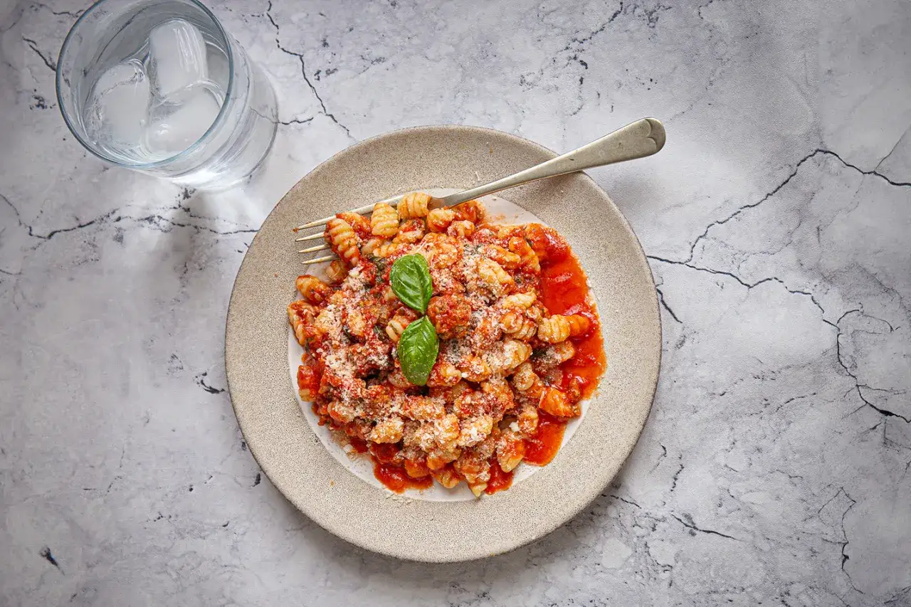 Gnocchetti Sardi Rezept: Herzhafte Pasta mit Tomatensauce, Parmesan und frischem Basilikum auf einem Teller, daneben ein Glas Wasser mit Eis.