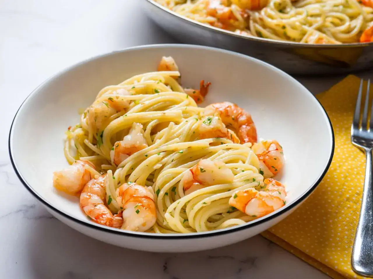 Ein Teller mit k&ouml;stlichem Spaghetti mit Garnelen, garniert mit Petersilie. Im Hintergrund eine Pfanne mit demselben Gericht.