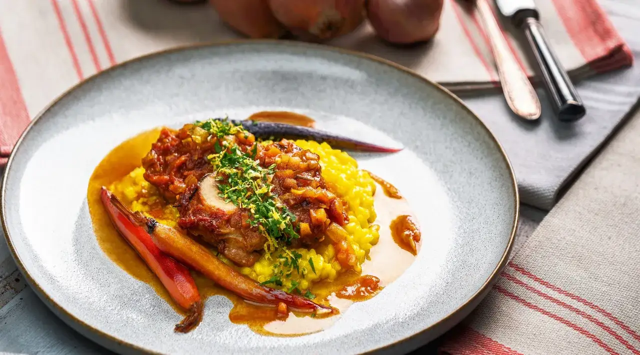 Ein Teller mit einem authentischen Rezept Ossobuco Original, serviert auf cremigem Safranrisotto, garniert mit frischen Kräutern und gerösteten Karotten.