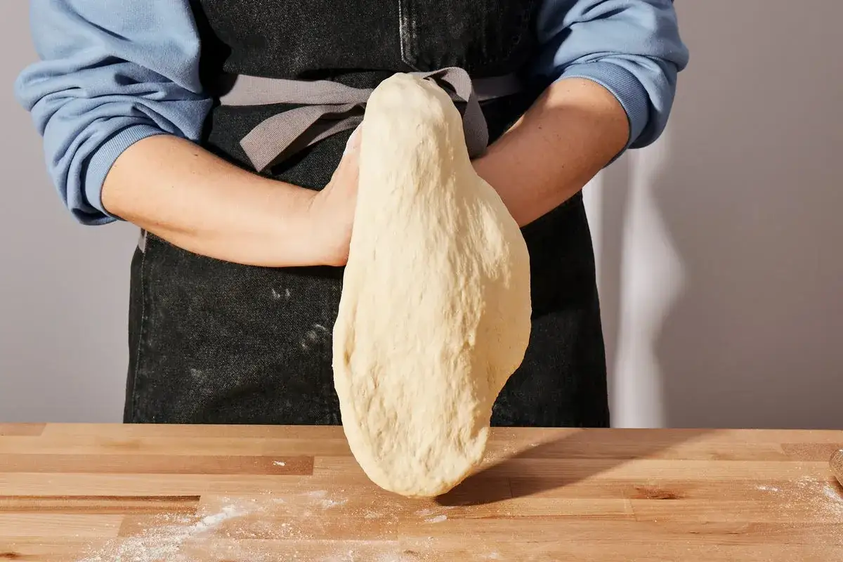 H&auml;nde formen einen gro&szlig;en, elastischen Teigling f&uuml;r Pizza. Der Teig ist hellgelb und liegt auf einem Holztisch.