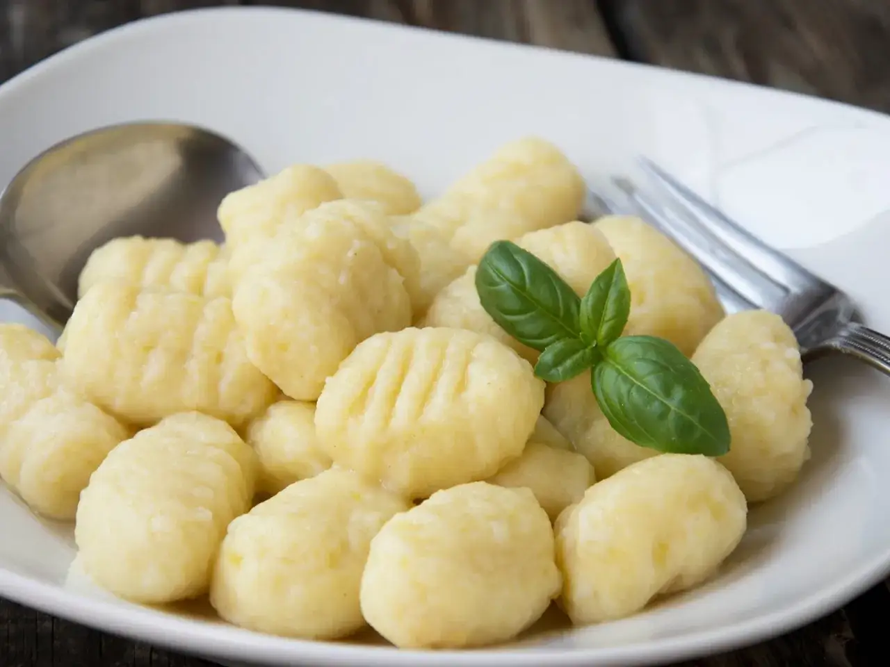 Ein Teller mit k&ouml;stlichen, italienischen Gnocchi, garniert mit frischem Basilikum. Daneben liegen L&ouml;ffel und Gabel.
