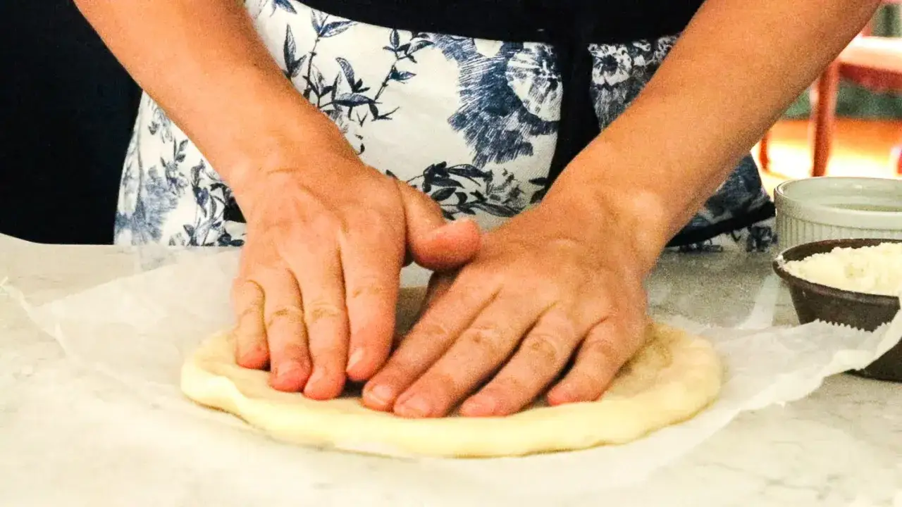 H&auml;nde formen einen runden pizzateig mit trockenhefe auf Backpapier. Daneben stehen Sch&uuml;sseln mit Zutaten.