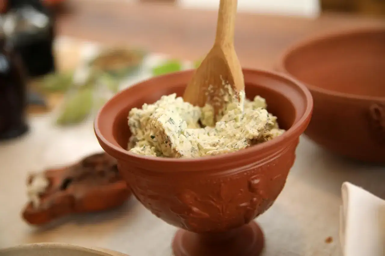 Ein rustikales Tongef&auml;&szlig; mit cremigem Moretum, einem Kr&auml;uter-K&auml;se-Dip, und einem Holzspatel. Perfekt f&uuml;r ein antikes Moretum Rezept.