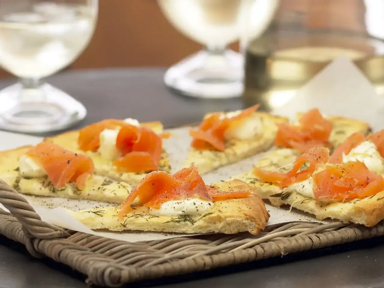 K&ouml;stliche kleine Pizza-St&uuml;cke mit Lachs und Spinat, serviert auf einem Korbtablett, mit Wei&szlig;wein im Hintergrund.