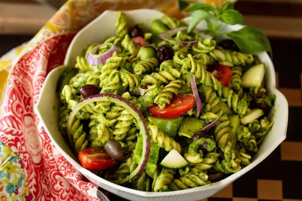 Ein köstlicher Nudelsalat mit Pesto, Tomaten, Oliven, Gurken und roten Zwiebeln, garniert mit Basilikum.