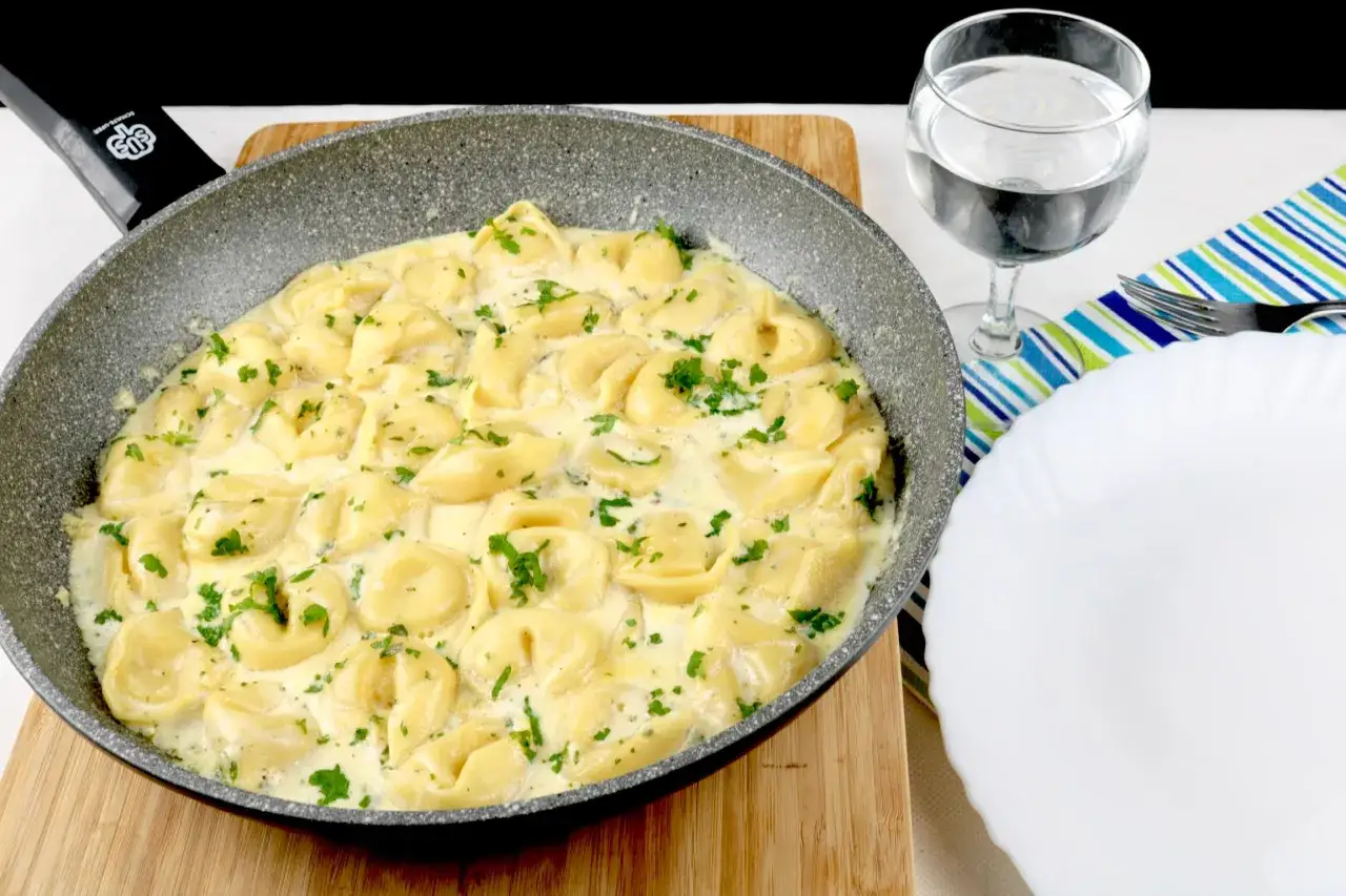 Cremige Tortellini in einer Pfanne, garniert mit Petersilie. Perfekt f&uuml;r schnelle Tortellini Rezepte. Daneben ein Glas Wasser und ein Teller.