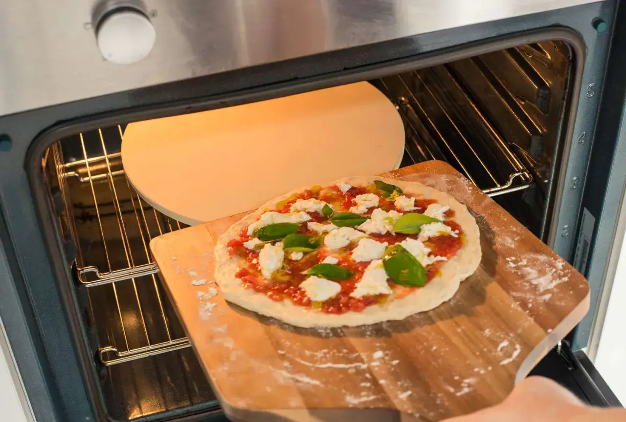 Eine Margherita-Pizza mit Mozzarella und Basilikum wird auf einem Pizzastein in den Ofen geschoben. Perfekt f&uuml;r Ihr n&auml;chstes Pizza backen Rezept!