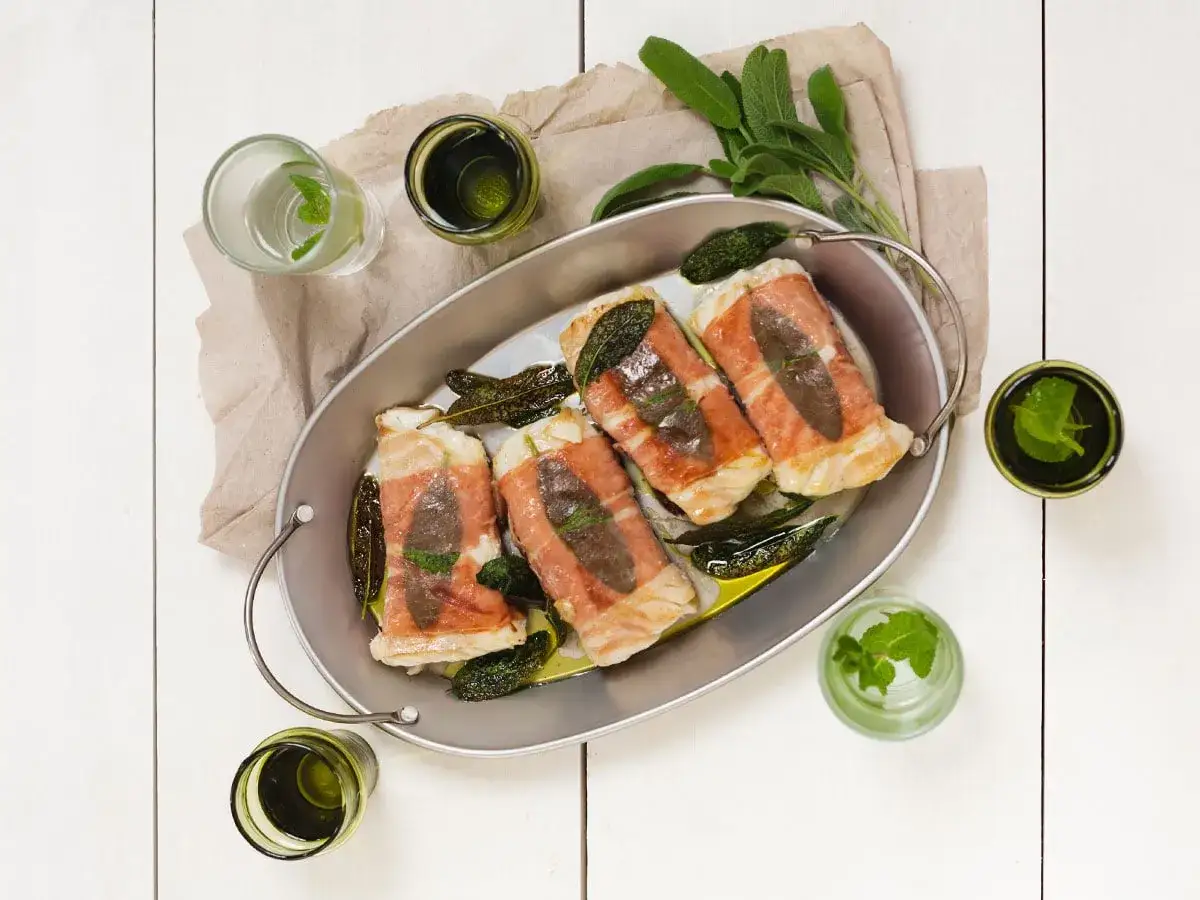 Kabeljau Saltimbocca in einer ovalen Schale, umwickelt mit Schinken und Salbei, serviert mit Kräutergetränken.