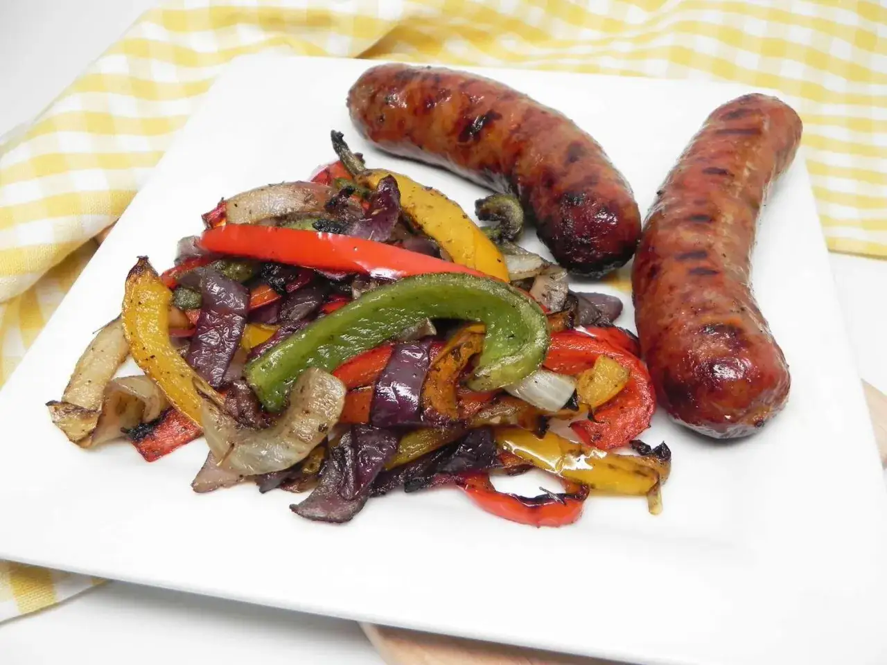 Zwei gegrillte Salsiccia liegen neben einem Haufen bunter, gegrillter Paprika und Zwiebeln auf einem wei&szlig;en Teller.