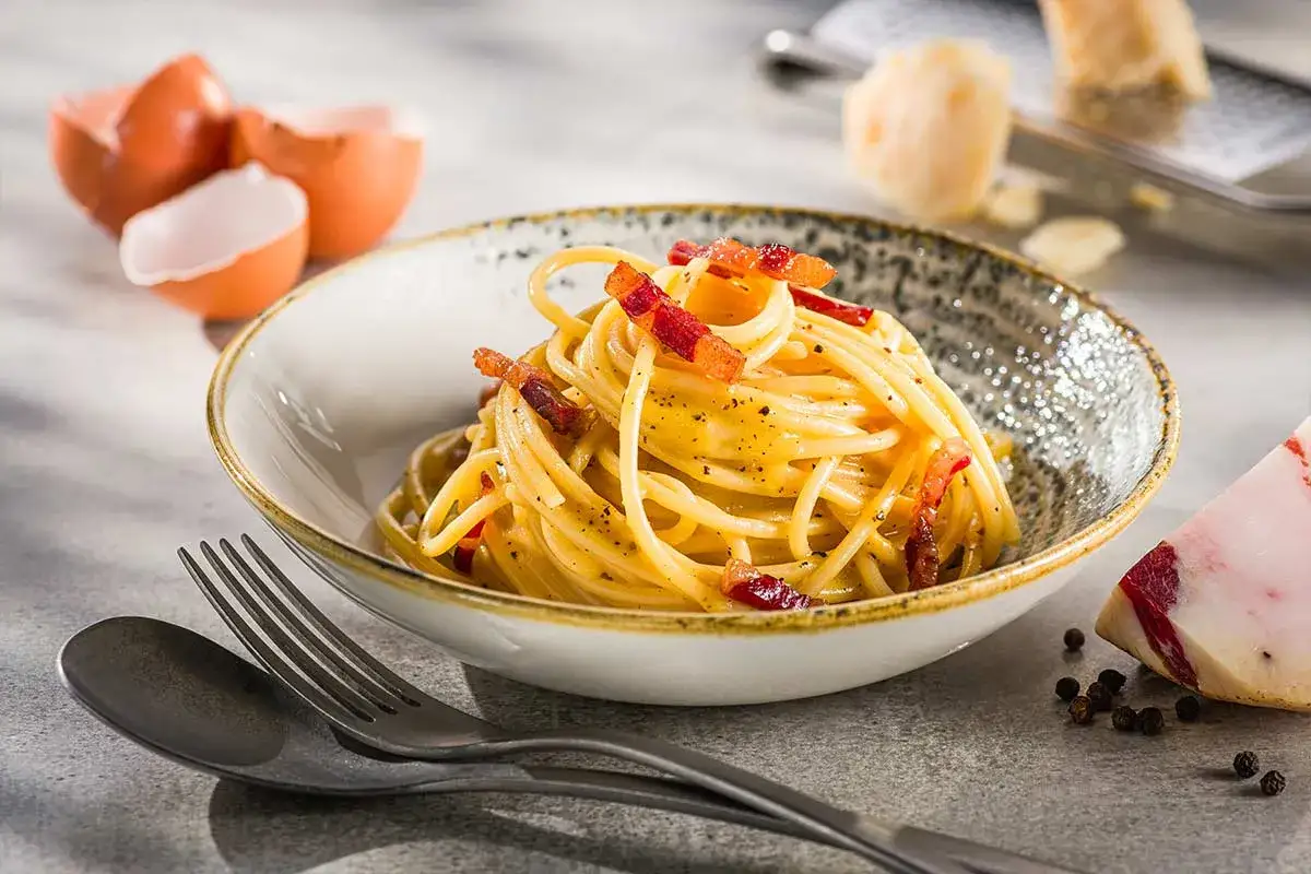 Eine Sch&uuml;ssel mit echter Carbonara, garniert mit Guanciale und schwarzem Pfeffer. Daneben liegen Eierschalen und ein St&uuml;ck Pecorino.