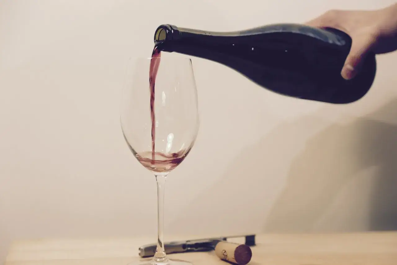 Ein Weinglas wird mit Rotwein gefüllt. Eine Hand hält die dunkle Flasche, während der Wein in einem dünnen Strahl ins Glas fließt.