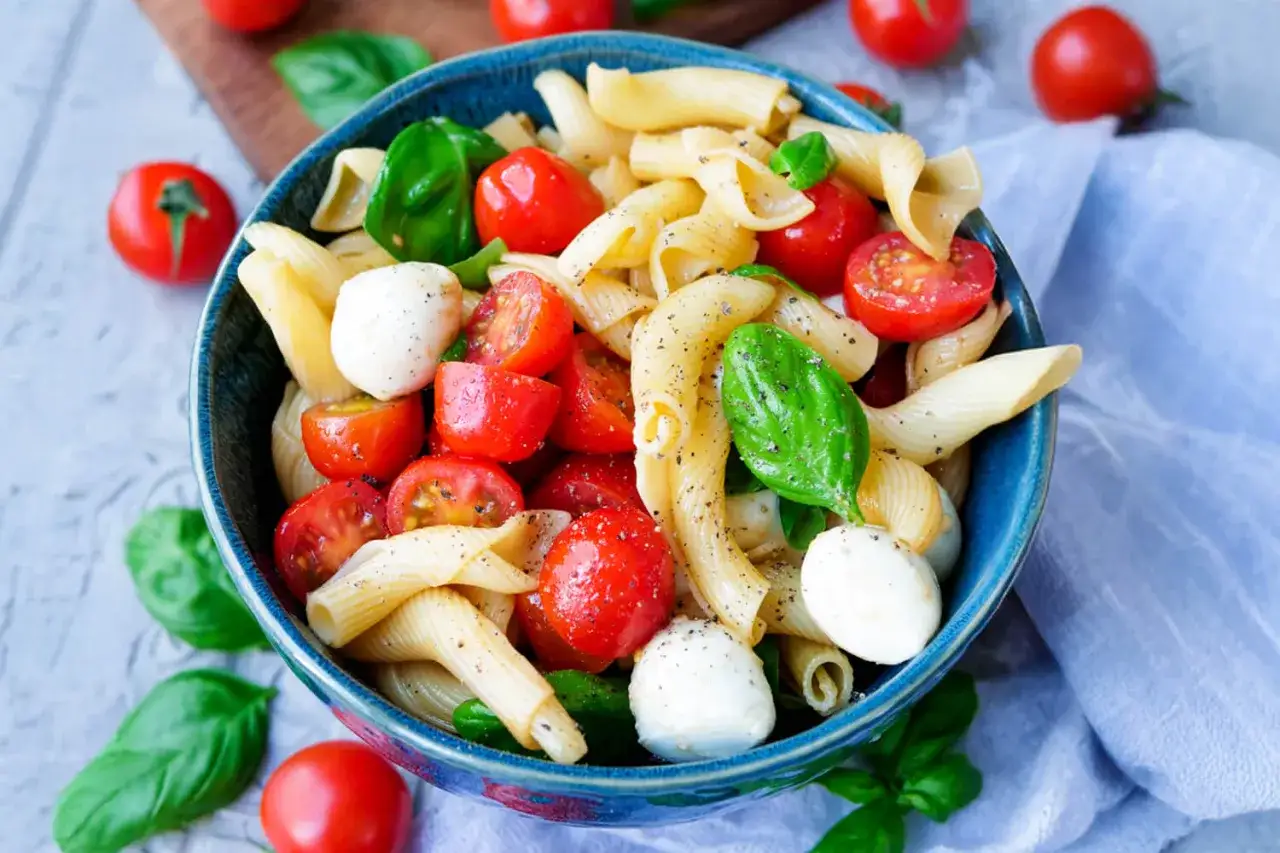 Ein bunter italienischer Nudelsalat mit Penne, Kirschtomaten, Mozzarella-Kugeln und frischem Basilikum, appetitlich in einer blauen Schüssel angerichtet.