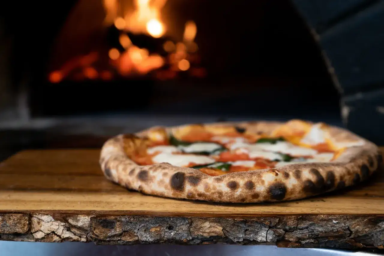 Eine frisch gebackene Pizza Margherita liegt auf einem Holzbrett vor einem gl&uuml;henden Ofen. So sieht eine Pizza aus, wenn sie perfekt zubereitet ist.