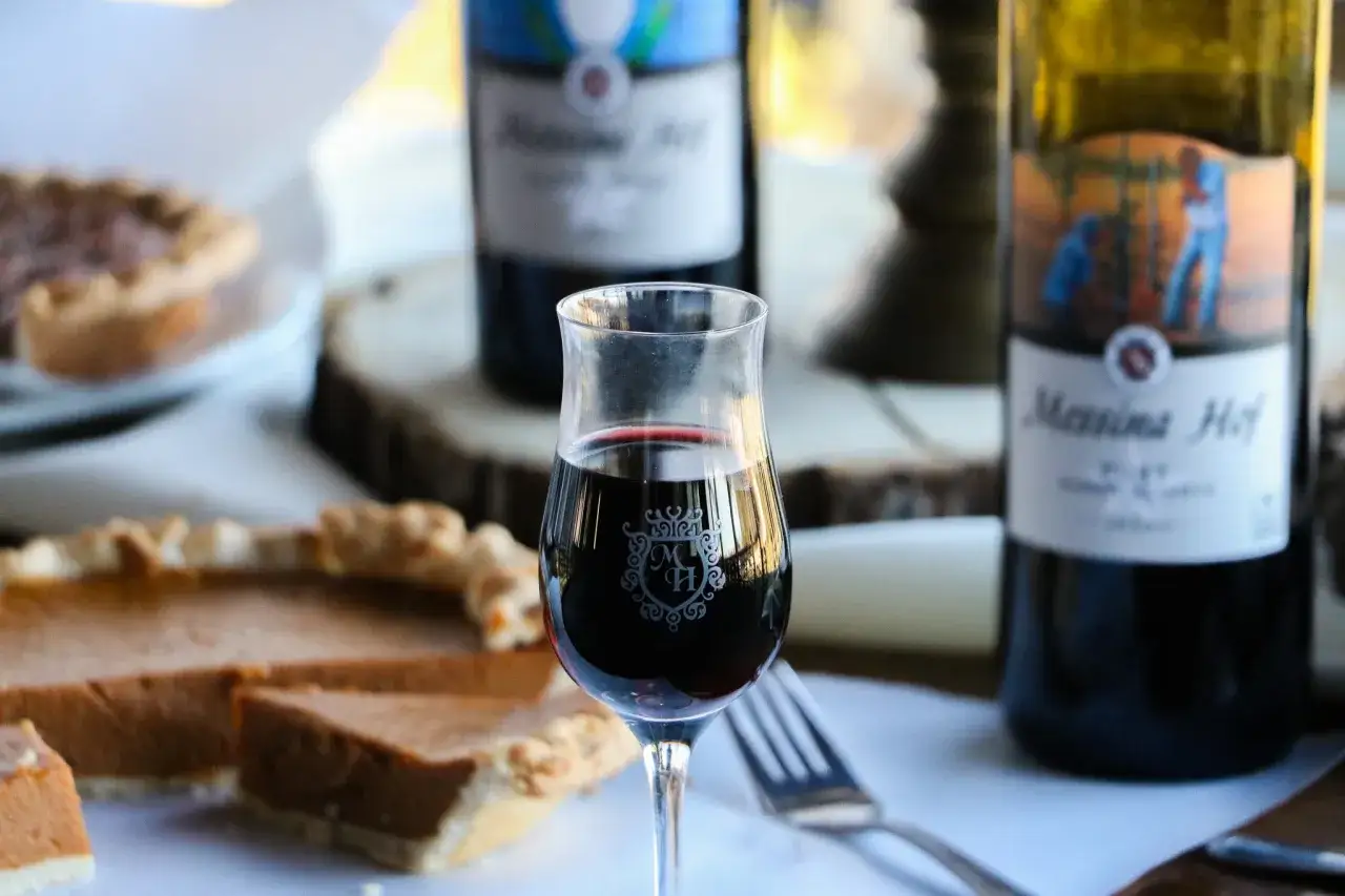Ein Glas Rotwein, dessen Trauben aus Vitis vinifera stammen, steht neben Kürbiskuchen und Messina Hof Weinflaschen.