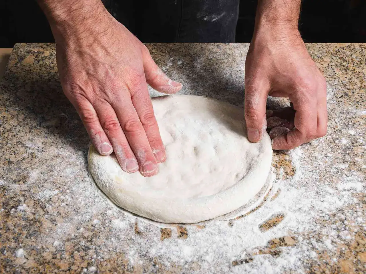 H&auml;nde formen einen Pizzateig auf einer bemehlten Arbeitsfl&auml;che. Der Teig wird gerade zu einer runden Pizza geformt.