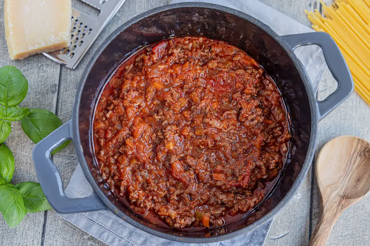 Herzhafte Bolognese-Sauce in einem Topf, umgeben von frischem Basilikum, Parmesan, Spaghetti und einem Holzlöffel. Alle Zutaten für eine köstliche Bolognese sind bereit.