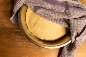 Frischer pizzateig für 4 Personen ruht in einer Schüssel unter einem karierten Tuch auf einem Holztisch.