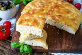 Frisch gebackener focaccia teig, aufgeschnitten und gestapelt, mit Tomaten und Oliven im Hintergrund.