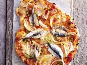 Eine rustikale Sardellen Pizza mit Zwiebeln und grünen Paprikastreifen, auf einem Holzbrett serviert.
