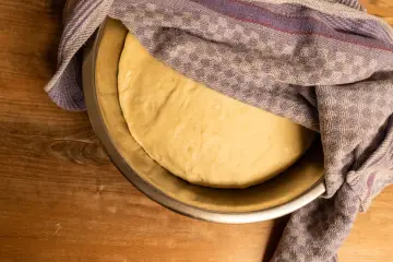 Frischer pizzateig für 4 Personen ruht in einer Schüssel unter einem karierten Tuch auf einem Holztisch.