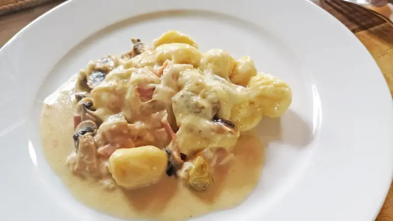 Gnocchi al forno mit cremiger Käsesauce, Pilzen und Schinken auf einem weißen Teller.