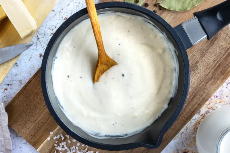 Cremige Béchamelsauce in einer Pfanne, bereit für Lasagne. Butter, Milch und Gewürze liegen daneben.