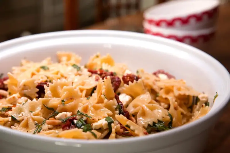 Ein köstlicher italienischer Nudelsalat mit Farfalle, getrockneten Tomaten, Feta und Kräutern in einer weißen Schüssel.
